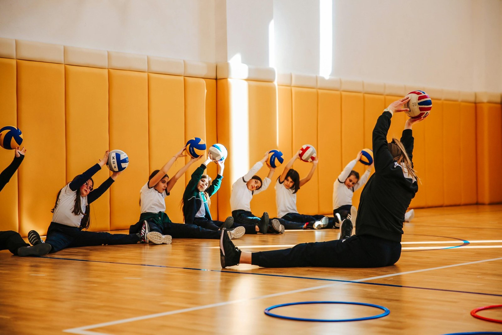 Group of children practicing volleyball stretches in a spacious indoor gym with a coach.