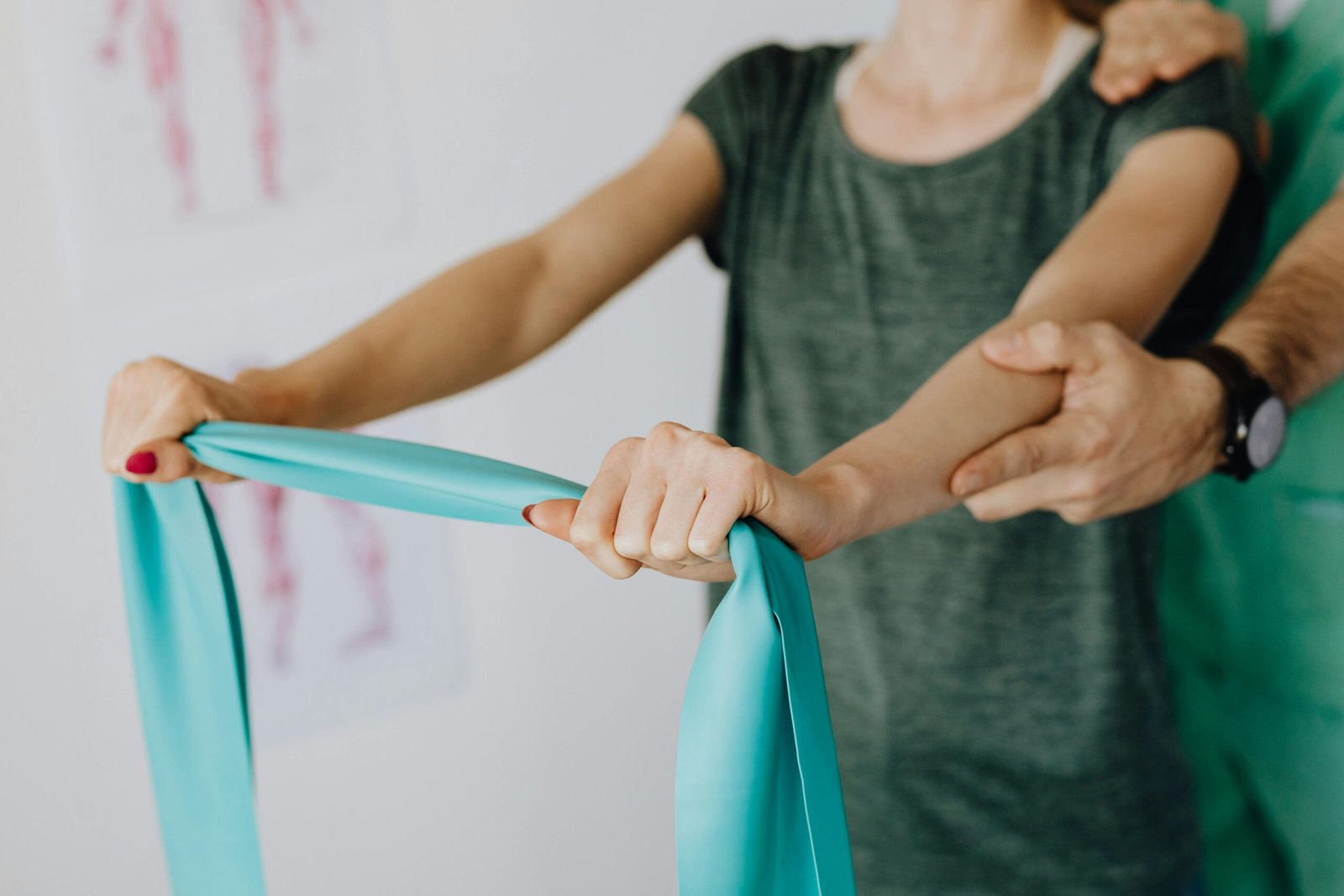 Crop unrecognizable orthopedist in uniform and wristwatch supporting shoulder and reached arm of faceless female patient with resistance band in doctor office